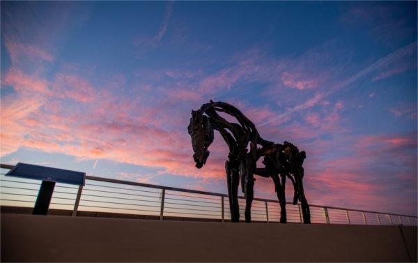 A horse sculpture is next to a railing with a pink sky in the background.