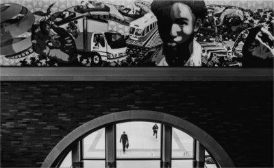 A mural is seen above doors of a building. People are seen walking outside through windows.