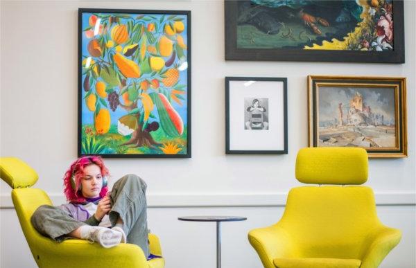 A person wearing headphones sits in a yellow chair. Several pieces of art on the wall in the background.